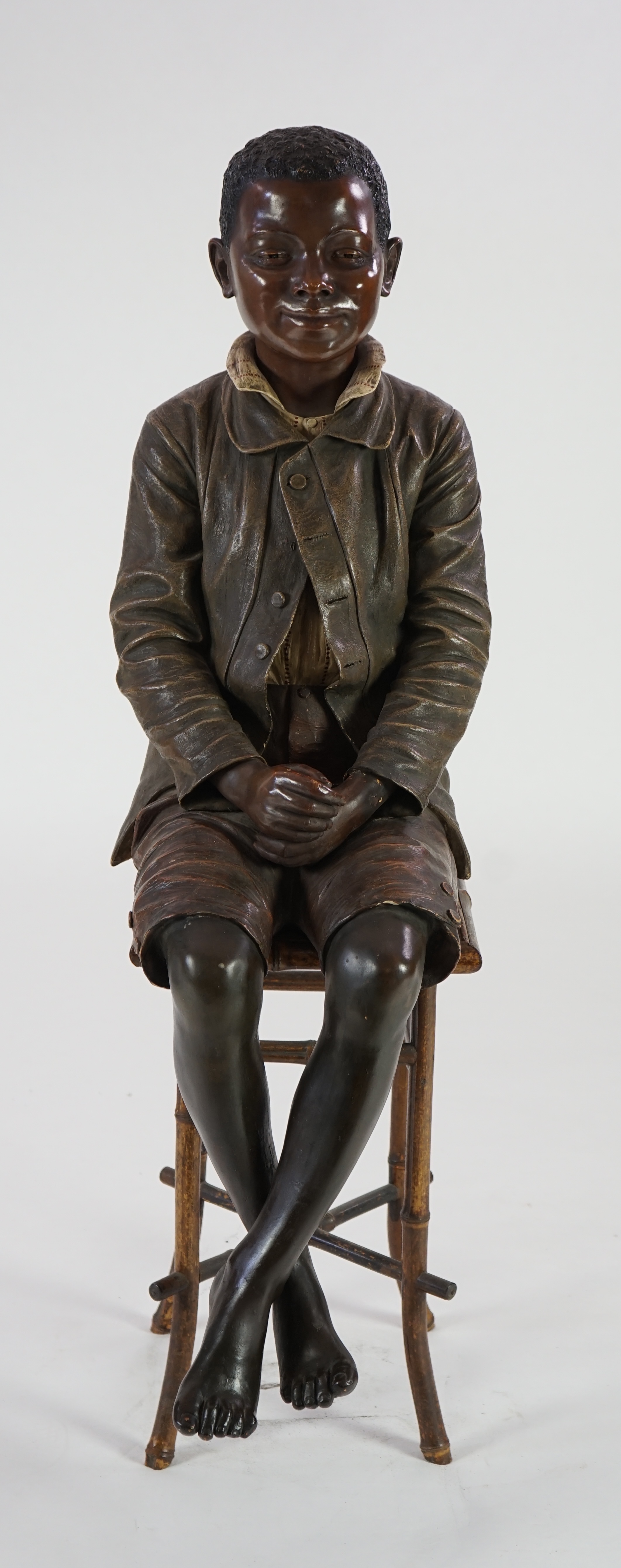 A late 19th century Friedrich Goldscheider life size cold painted terracotta figure of a black boy, seated upon a bamboo and wicker stool, impressed, 33cm wide, overall 116cm high. Condition - poor to fair, feet damaged